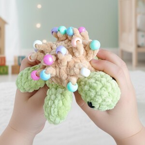 May include: A crocheted turtle toy in tan and green, decorated with colourful beads. The turtle's body is a textured tan colour, with green flipper-like appendages. The beads are in various pastel colours, adding a playful touch to the toy.