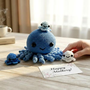 Pode incluir: Um polvo azul de crochê feito à mão com um polvo menor em cima e dois polvos pequenos adicionais. Um cartão com o texto "Happy Mothery!" também está presente.