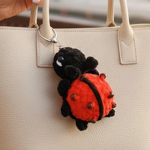 Può includere: Un portachiavi a forma di coccinella fatto a mano. La coccinella ha un corpo rosso con pois neri e una testa e zampe nere. Il portachiavi è attaccato a un anello d'argento ed è appeso a una borsa beige.