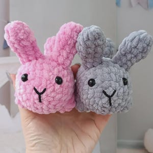 May include: Two plush bunny toys, one pink and one grey, are held in a hand. Each bunny has black eyes and a black stitched mouth. The bunnies are made of a soft, textured material.