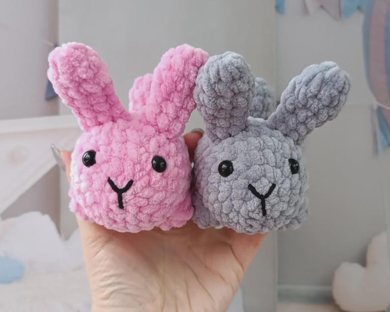 May include: Two plush bunny toys, one pink and one grey, are held in a hand. Each bunny has black eyes and a black stitched mouth. The bunnies are made of a soft, textured material.