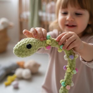 May include: A crocheted green snake toy with a black eye and colourful beads. The snake is held by a child. The toy is made of soft yarn and has a textured appearance. The beads are pastel colours.