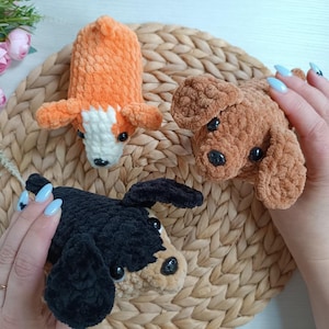 May include: Three handmade crochet dog toys in orange, brown, and black. The orange dog has white markings, the brown dog has black eyes, and the black dog has tan markings. The toys are made of soft, textured yarn.