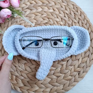 May include: A light blue crocheted elephant-shaped glasses holder with a pair of glasses resting inside.