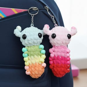 May include: Two handmade crochet keychains. One is light blue, the other is pink. Both have black eyes, small horns, and a beaded body in a gradient of colors. They are attached to silver keyrings and hang against a navy blue backpack.
