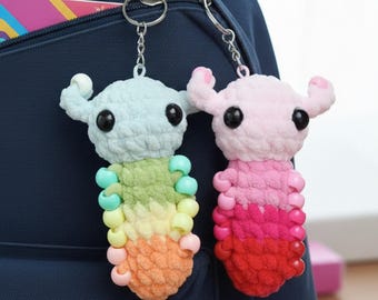 May include: Two handmade crochet keychains. One is light blue, the other is pink. Both have black eyes, small horns, and a beaded body in a gradient of colors. They are attached to silver keyrings and hang against a navy blue backpack.
