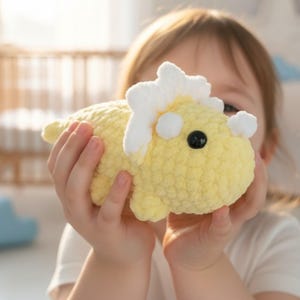 May include: A handmade, yellow crocheted dinosaur toy with white accents on its head and back. The dinosaur has a black button eye and is held in a child's hands. The toy is soft and plush.