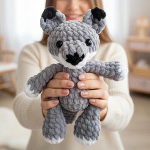 May include: A handmade crochet plush toy shaped like a fox, crafted from thick, textured grey yarn. The fox features black-tipped ears, a black nose, white accents on the muzzle and paws, and two round, glossy black plastic eyes.