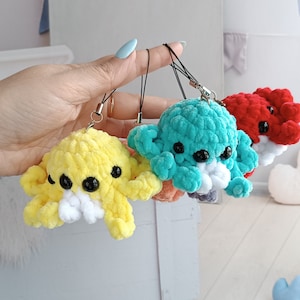 May include: Four crocheted octopus plushies in different colors: yellow, teal, pink, and red. Each octopus has black eyes and a white crocheted mouth. The plushies are attached to keychains.