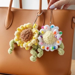 May include: Two handmade crochet sunflower keychains. One sunflower is yellow and brown, the other is white and yellow with colorful beads. The keychains are attached to a brown leather tote bag.