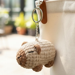 May include: A crocheted capybara keyring in beige and brown, with closed eyes, attached to a silver chain and keyring. The keyring is hanging from a beige tote bag.