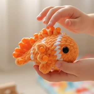 May include: A handmade orange and white crocheted fish toy with a black eye. The fish has orange fins and a white stripe pattern on its body. The toy is held in a person's hands.