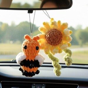 May include: Crocheted car charms featuring a bee and a sunflower. The bee is orange and black with white wings. The sunflower is yellow and brown with a green stem. Both charms are hanging from a rearview mirror.