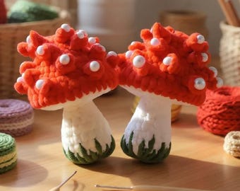 May include: Two handmade crochet mushrooms with red caps dotted with white beads. The stems are white with green accents at the base. The mushrooms are displayed on a wooden surface, suggesting a craft or home decor theme.