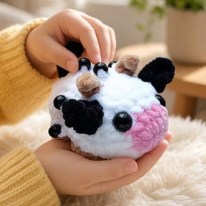 May include: A small, round, crocheted plush toy shaped like a cow. The body is white with black patches, a pink snout, and small brown accents. It features shiny black plastic eyes and small black beads attached to the top, crafted from soft, textured yarn.