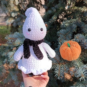 Può includere: Un peluche a forma di fantasma lavorato all'uncinetto in bianco con una sciarpa nera, occhi di sicurezza neri e guance ricamate in rosa, accompagnato da una piccola zucca all'uncinetto arancione con un picciolo verde, in filato morbido.