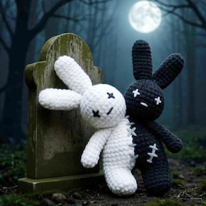 May include: Two crochet bunny dolls, one white and one black, stand side-by-side in front of a tombstone. The white bunny has black cross-stitch eyes and mouth, while the black bunny has white cross-stitch details. The background features a full moon and a dark forest.