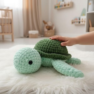 Puede incluir: Un peluche de tortuga hecho a mano con ganchillo en tonos verdes. La tortuga tiene una cabeza y un caparazón redondeados, con un botón negro como ojo. El peluche está sobre una superficie blanca y esponjosa. La mano de un niño toca suavemente el caparazón de la tortuga.