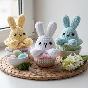 May include: Three crocheted bunny figures in yellow, white, and blue, each in a pastel cupcake base. The bunnies have black eyes and a stitched "Y" mouth. A small bowl of pastel Easter eggs is placed nearby. The scene is set for Easter.