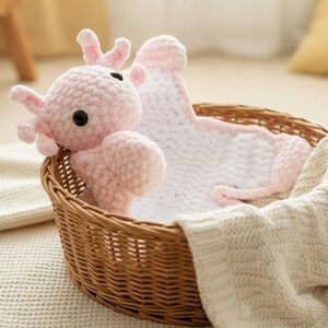 May include: A handmade pink crochet elephant toy with black eyes and a matching blanket, nestled in a woven wicker basket. The blanket is white with pink trim. A cream-colored blanket is draped over the basket.