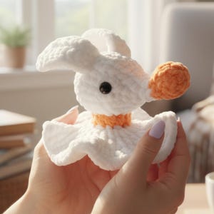 Pode incluir: Um ganso branco de crochê feito à mão com bico e gola laranja. O ganso tem olhos pretos e uma saia com babados. O brinquedo de pelúcia é segurado nas mãos de uma pessoa. Um brinquedo macio e caprichoso.