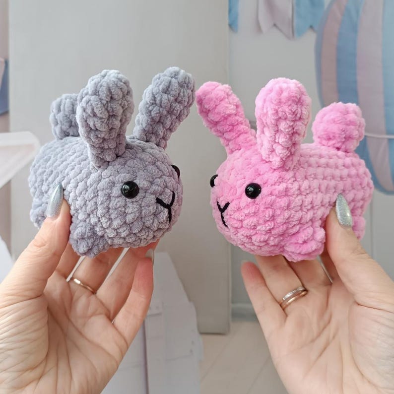May include: Two handmade crochet bunnies, one grey and one pink, are held up. Each bunny has black eyes and a stitched mouth. The bunnies are soft and plush, with long ears and small tails. The background is blurred.