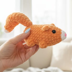 May include: An orange crocheted hamster toy with a curved tail and small ears. The hamster has a white nose and a pink tip, with black eyes. The toy is held in a hand.