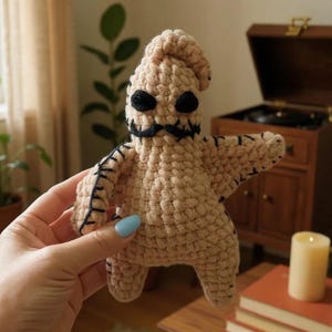 May include: A tan crocheted Oogie Boogie doll with black eyes, a black mustache, and black stitching. The doll has a star-like shape with arms outstretched. The doll is handmade and has a textured appearance.