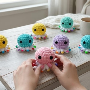 May include: A collection of small, round, crocheted octopus toys in various pastel colors including yellow, teal, lavender, and pink. Each octopus features black plastic eyes and white tentacles with multicolored tips, arranged on a wooden surface.