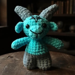 May include: A handmade crochet plush toy in the shape of a goat. The goat is turquoise with gray horns and legs. A black pentagram is stitched on the forehead, and the eyes are black. The toy is sitting on a wooden surface.