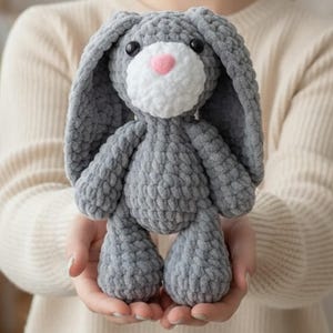 May include: A plush toy rabbit made of thick gray yarn with a white muzzle and a small pink nose. The stuffed animal features long, floppy ears and two round black eyes. The texture of the yarn is chunky and soft, creating a rounded, tactile form.