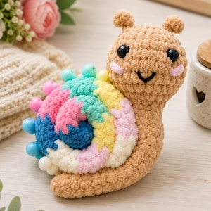 May include: A crocheted snail toy with a tan body and a multi-coloured spiral shell. The shell features segments of blue, pink, yellow, and white yarn, accented with small, round, colourful plastic beads along the outer edge. The snail has black eyes.