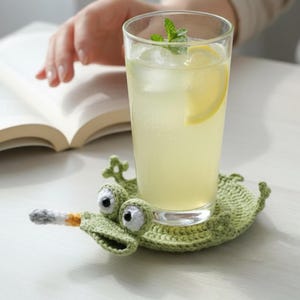 Peut inclure: Un dessous de verre au crochet vert clair en forme de grenouille avec de grands yeux blancs et noirs et une petite protubérance grise et orange ressemblant à une cigarette, supportant un verre transparent rempli d'une boisson jaune.