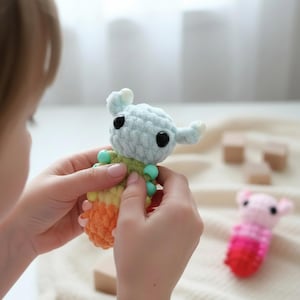 May include: A handmade crochet toy in the shape of a small alien creature. The toy is light blue with large black eyes, small ears, and a colorful body with green, yellow, and orange sections. The toy is held in a person's hands.