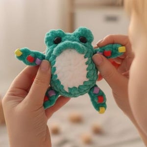 May include: A teal crochet frog toy with white, yellow, red, and purple accents. The frog has black eyes and is made of soft yarn. The frog's arms and legs have colourful details. A charming and unique handmade toy.