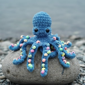 Puede incluir: Un pulpo azul tejido a mano con ojos negros y cuentas coloridas en sus tentáculos. El peluche está sentado sobre una roca gris, con un fondo borroso de agua y guijarros.
