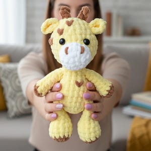 May include: A handmade, yellow crochet giraffe plush toy with brown accents on the hooves, ears, and horns. The giraffe has a white muzzle, black eyes, and heart-shaped markings. The soft toy is held up in front of a blurred background.