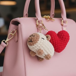 Könnte beinhalten: Eine rosa Lederhandtasche mit goldfarbener Hardware. Ein Schlüsselanhänger mit einem gehäkelten Schaf und einem roten Herzanhänger hängt am Griff. Das Schaf ist weiß und braun, und das Herz ist rot.