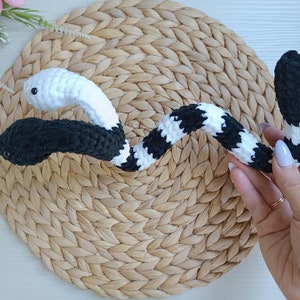 May include: A black and white crocheted snake toy with a black head and a white tail. The snake is curled up on a woven mat.