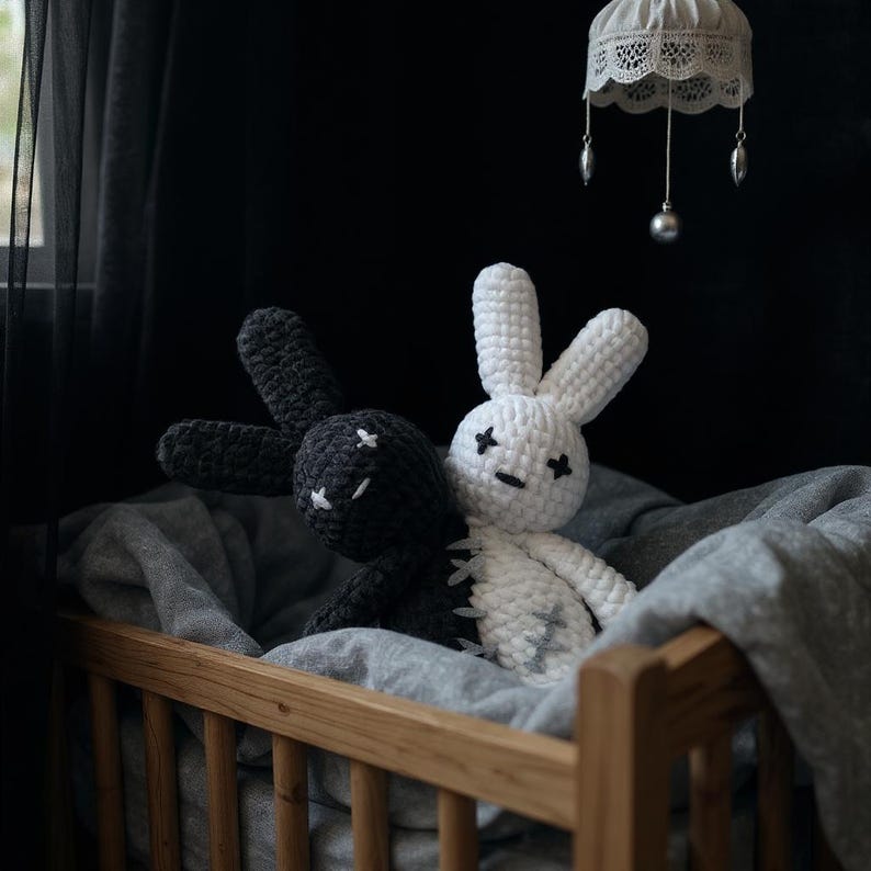 Pode incluir: Duas bonecas de coelho de croch&ecirc; feitas &agrave; m&atilde;o, uma preta e outra branca, repousam em um ber&ccedil;o de madeira. Os coelhos t&ecirc;m tra&ccedil;os faciais costurados e est&atilde;o colocados em um fundo escuro. Um cobertor cinza e uma lumin&aacute;ria suspensa decorativa tamb&eacute;m s&atilde;o vis&iacute;veis.