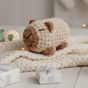 Może przedstawiać: Mała, ręcznie robiona pluszowa zabawka na szydełku w kształcie kapibary. Korpus wykonany jest z grubej, teksturowanej przędzy w kolorze kremowym, natomiast pyszczek, uszy i stopy z kontrastującej jasnobrązowej przędzy. Posiada proste, czarne oczy.