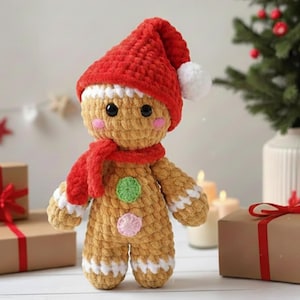 Könnte beinhalten: Eine beige gehäkelte Lebkuchenpuppe mit einer roten Mütze mit weißem Bommel und einem passenden roten Schal. Die Puppe hat schwarze Knopfaugen, rosa Wangen, einen grünen runden Bauchnabel, einen rosa runden Bauchnabel und weiße Verzierungen.