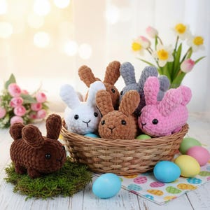 May include: A selection of handmade crochet bunny rabbits in a wicker basket and on moss. The bunnies are in various colours, including white, brown, grey, and pink. Pastel-coloured Easter eggs are scattered around the basket.