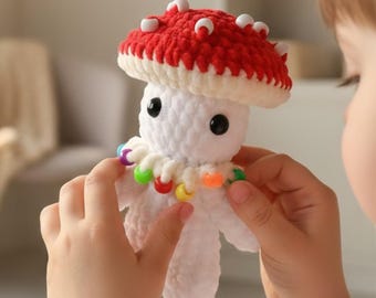 May include: A handmade crochet mushroom toy with a red cap, white spots, and a white stem. The mushroom has black eyes and a colorful beaded necklace. The toy is being held by a person.
