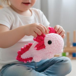 Puede incluir: Un peluche de axolotl rosa y fucsia hecho a mano con ganchillo. El juguete tiene una cabeza redondeada, un cuerpo largo y branquias detalladas. El axolotl está siendo sostenido por una persona que lleva una camiseta blanca y vaqueros azules.