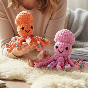 Puede incluir: Dos juguetes de pulpo hechos a mano con ganchillo. Uno es naranja, el otro rosa. Ambos tienen ojos negros y caras sonrientes. Los tentáculos del pulpo están decorados con cuentas de colores. Los juguetes están hechos de hilo suave.