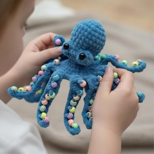 May include: A handmade blue crochet octopus toy with black eyes and colorful beads on its tentacles. The plush toy is held by a person, showcasing the detailed craftsmanship and playful design. A soft, textured, and whimsical creation.