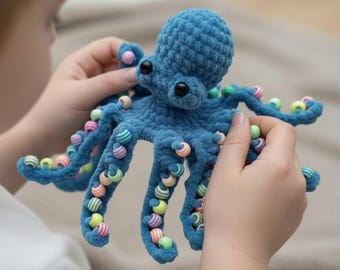 May include: A handmade blue crochet octopus toy with black eyes and colorful beads on its tentacles. The plush toy is held by a person, showcasing the detailed craftsmanship and playful design. A soft, textured, and whimsical creation.