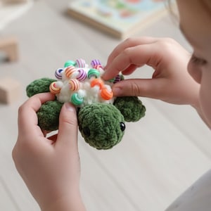May include: A handmade green crochet turtle toy with a white shell adorned with colorful beads. The turtle has black eyes and is being held by a person. The toy is designed for children.