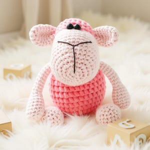 May include: A handcrafted pink and white crochet sheep toy with black eyes and a black stitched face. The sheep has a round body, four limbs, and large ears. The toy is sitting on a white fluffy surface.